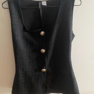 Svart topp  - Stilren svart väst från H&M med markerad struktur och snygga guldiga knappar framtill. Västen har en rak passform och är ärmlös, perfekt att styla över en skjorta eller t-shirt för en trendig look. fin figurnära topp, ser ut som en kostym topp och sitter väldigt fint på