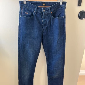 Mörkblå jeans från BOSS - Snygga mörkblå jeans från BOSS med klassisk femficksdesign och knappgylf. Jeansen har raka ben och subtila slitningar för en modern look. Perfekta till sneakers eller boots och passar dig som gillar stilrena och tidlösa plagg.