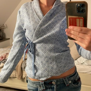 Blå omlottkofta med mönster från Odd Molly  - Supersöt blå och vit omlottkofta med knyt i sidan. Koftan har ett diskret mönster och är stickad i mjukt material. Perfekt för dig som gillar en chill men ändå stilren look. Långärmad och passar till jeans eller kjol.