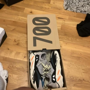 Adidas Yeezy 700 Wave Runner sneakers - Säljer ett par Adidas Yeezy 700 Wave Runner sneakers med chunky sula. Skorna har grå mesh och mocka, svarta och blå detaljer samt orange accenter på sulan. Klassisk dad shoe-vibe med snörning och rund tå. Perfekta för dig som vill sticka ut med en ikonisk modell. Men liten defekt på ena skon och lite smutsiga
