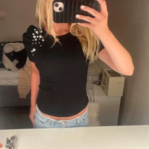 Svart ribbad topp med pärlor - Svart ribbad topp med korta puffärmar och snygga pärldetaljer på ena axeln. Tight passform som framhäver figuren och ger en trendig look. Perfekt att styla med jeans eller kjol för en cool outfit.