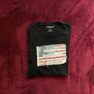Svart t-shirt Ralph Lauren flaggtryck - Svart t-shirt från Ralph Lauren Denim & Supply med ett stort vintageinspirerat amerikanskt flaggtryck på bröstet. Klassisk rund hals och korta ärmar. Mjuk bomullskvalitet som känns skön mot huden. Perfekt för en avslappnad och cool stil.