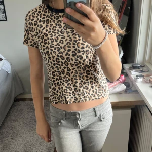 Leopardmönstrad topp från H&M  - Säljer en kortärmad topp från H&M Divided i storlek XS med leopardmönster i beige, brunt och svart. Toppen har en svart ribbad krage och är croppad i modellen. Perfekt för dig som vill sticka ut med ett djurmönstrat plagg.