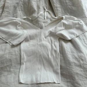 Vit  topp med puffärm och knyt - Supersöt vit ribbad topp med puffiga korta ärmar och v-ringning. Toppen har knytband i nacken och är figurnära i modellen. 