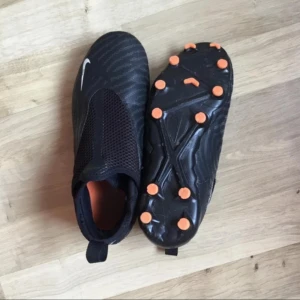 Svarta Nike fotbollsskor med orange dobbar - Säljer ett par svarta Nike fotbollsskor med orange dobbar och diskret mönster på ovandelen. Skorna har en strömlinjeformad design utan synlig snörning och meshpanel över vristen för extra ventilation. Perfekta för dig som vill ha stilrena och funktionella skor på planen.