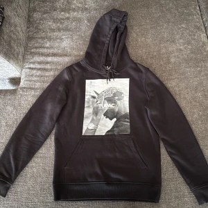 Svart Tupac hoodie Mister Tee S - Svart hoodie från Mister Tee x Artists med stor Tupac-print på bröstet. Tröjan har huva med snörning, känguruficka och är tillverkad i mjuk bomulls- och polyestermix. Perfekt för dig som gillar streetwear och vill ha en ikonisk look.