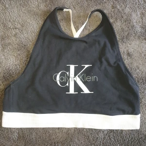 Svart bh/topp från Calvin Klein - Svart bh från Calvin Klein med vitt CK-tryck framtill. Toppen har vita justerbara axelband som korsas i ryggen och en bred, ribbad vit kant nedtill. Perfekt för en sportig och stilren look.