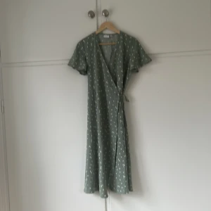 Grön blommig omlottklänning från Vila - Supersöt grön omlottklänning från Vila med små vita blommor. Klänningen har korta ärmar, knyt i midjan och faller i en längre modell. Orginalpris 499 säljer för 300kr