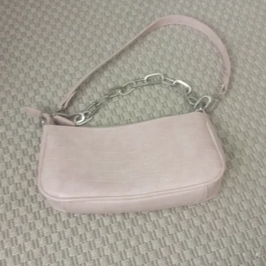 Ljusrosa axelväska från Gina Tricot - Säljer en ljusrosa axelväska från Gina Tricot med croco-mönstrad yta och silvrig kedjedetalj. Väskan har dragkedja upptill och är i syntetmaterial. Perfekt för dig som vill ha en trendig och stilren accessoar.