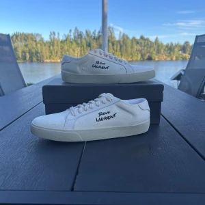 Vita Saint Laurent sneakers i canvas - Snygga vita sneakers från Saint Laurent med handskriven logga på sidan. Skorna är i canvas med vita snören och platt sula. Perfekt minimalistisk design som passar till allt. Kommer med originalkartong och dustbag. Tar även byten mot maison margiela gats skor. Hör av dig vid minsta lilla fundering.