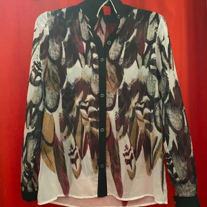 Mönstrad blus med fjädertryck - Snygg blus med fjäderinspirerat mönster i svart, beige, brun och vinröd. Långärmad modell med svarta knappar och svarta detaljer längs knappslån och vid ärmslut. Tunn och luftig, perfekt för dig som gillar unika prints.