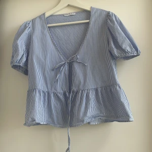 Blårandig blus med knyt från Bershka! - Supersöt blårandig blus från Bershka med v-ringning och knyt framtill. Puffiga korta ärmar med resår och volang nedtill. Säljer denna då den tyvärr inte har kommit till användning, har endast använts kanske 2-3 gånger💙🤍                                 (Självklart kan man knyta det nedre också, glömde göra det när jag tog bilden😊)