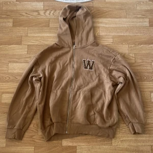 Brun hoodie med dragkedja från H&M - Säljer en brun hoodie från H&M i relaxed fit med dragkedja och huva. Tröjan har ett broderat W på bröstet och är fodrad med mjuk fleece på insidan. Perfekt för chill dagar och enkel att matcha med jeans eller joggers.