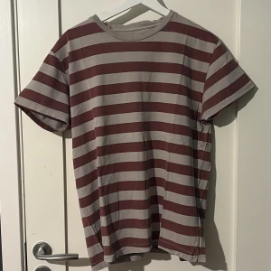 Randig t-shirt i vinrött och grått - Snygg t-shirt med breda horisontella ränder i vinrött och ljusgrått. Klassisk rund halsringning och korta ärmar. På ryggen finns ett tryck av en person med gitarr i rött. Perfekt för dig som gillar en avslappnad och cool stil.