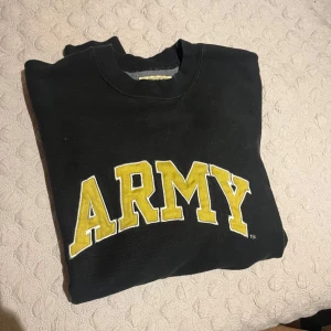 Svart sweatshirt med ARMY-tryck VINTAGE - Cool svart sweatshirt med stort gult ARMY-tryck på bröstet. Klassisk rund halsringning och långa ärmar. Perfekt för dig som gillar college-vibe och streetstyle. Mjuk och skön, passar till jeans eller joggers.