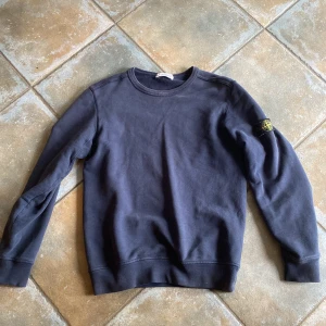 Mörkblå sweatshirt från Stone Island - Snygg mörkblå sweatshirt från Stone Island med klassisk rund hals och ikonisk logga på vänster ärm. Tröjan är i mjuk bomull och har långa ärmar samt ribbade muddar vid ärmslut och nederkant. Perfekt för en clean och stilren look. Storlek XS/junior