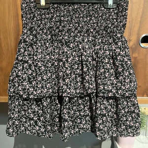 Blommig volangkjol från KappAhl - Supersöt kort volangkjol från KappAhl med svart botten och små ljusrosa blommor. Kjolen har smock i midjan och flera lager volanger som ger en flowy känsla. Perfekt för dig som gillar en feminin och trendig stil.