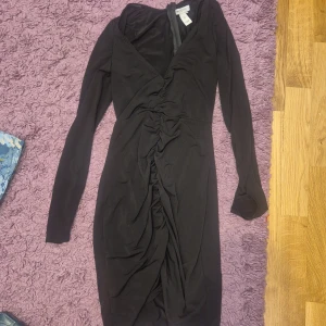 Svart långklänning från NLY One - Snygg svart långklänning från NLY One med lång ärm och v-ringad hals. Klänningen har en cool rynkad detalj framtill och dragkedja i ryggen. Perfekt för dig som vill ha en stilren och elegant look.