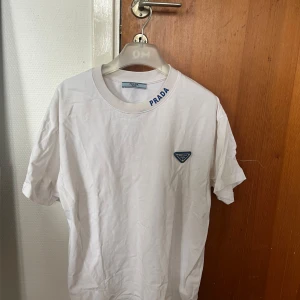 Prada T-shirt - Vit t-shirt från Prada i mjuk bomull med klassisk passform. T-shirten har blå Prada-logga på bröstet och tryckt Prada-text vid halsen. Enkel och clean design som ger en lyxig känsla till din outfit. Inget kvitto och därav priset