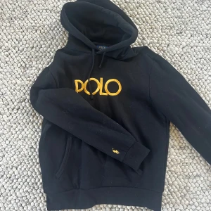 Svart hoodie oanvänd från Polo Ralph Lauren  - Svart hoodie från Polo Ralph Lauren med stor gul POLO-logga broderad på bröstet och liten gul polospelare på ärmen. Tröjan har huva med snörning och en klassisk känguruficka framtill. Perfekt för en chill och stilren look.