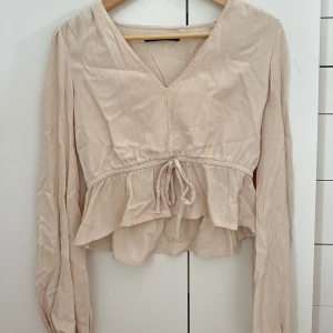 Beige blus med volang och knyt - Supersöt beige blus med v-ringning, långa ärmar och volang nedtill. Väldigt bra skick💖hör av er vid frågor om pris eller annat!