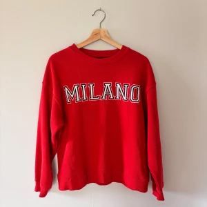 Röd Milano sweatshirt i bomull - Röd sweatshirt med trycket 'MILANO' i vitt och svart på bröstet. Tröjan har rund halsringning, långa ärmar och en relaxed passform. Köpt för 299 kr, använd 1-3 ggr!