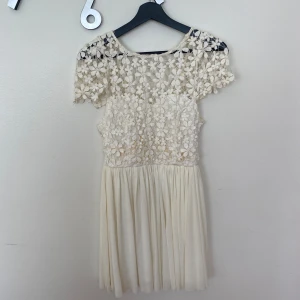 Vit spetsklänning från Xenia Boutique - Superfin kort vit klänning från Xenia Boutique med blommig spets upptill och plisserad kjol. Klänningen har korta ärmar, rund hals och dragkedja i ryggen. Använd fåtal gånger.