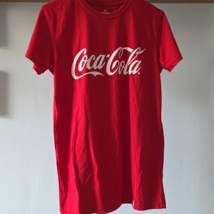 Röd Coca-Cola t-shirt i bomull/polyester - Röd t-shirt med klassiskt Coca-Cola-tryck i vitt framtill och liten flaska bak i nacken. Skön passform, rund hals och korta ärmar. Tillverkad av 70% återvunnen polyester och 30% Fairtrade-certifierad bomull. Perfekt för dig som gillar retro vibes och ikoniska loggor. Pris kan diskuteras :) 