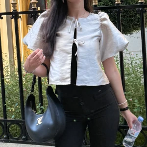 Champagnefärgad blus med puffärm - Säljer en champagnefärgad blus med korta puffärmar och croppad passform. Blusen har två söta knytband framtill och är tillverkad i ett glansigt material som ger en lyxig känsla. Perfekt för dig som gillar statement-ärmar och trendiga detaljer.