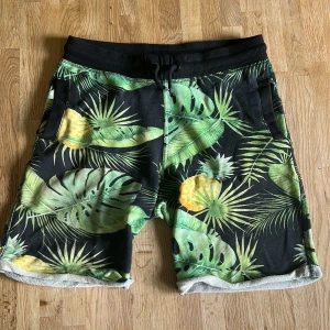 Svarta shorts med tropiskt mönster - Shorts med svart botten och tropiskt mönster med gröna palmblad och gula ananaser. De har elastisk midja med snörning och fickor både fram och bak. Materialet känns som mjuk bomull, perfekt för chill dagar. Tropisk vibe och riktigt sköna. Notera fläcken (se bild 4)