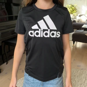 Svart Adidas t-shirt med stor logga - Svart t-shirt från Adidas med stor vit logga framtill. Klassisk rund halsringning och korta ärmar. Tillverkad i ett lätt och sportigt polyestermaterial som andas, perfekt för träning eller chill. Snygg och enkel design som funkar till jeans eller tights.