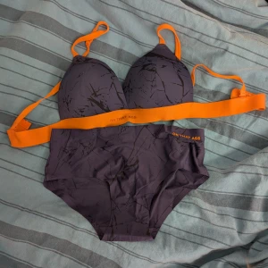 Mörkblå underklädesset från ON THAT ASS - Snyggt underklädesset från ON THAT ASS i mörkblå med svarta abstrakta mönster och coola orangea band. Toppen har vadderade kupor och breda axelband, medan trosan har en bekväm passform/hipster. 