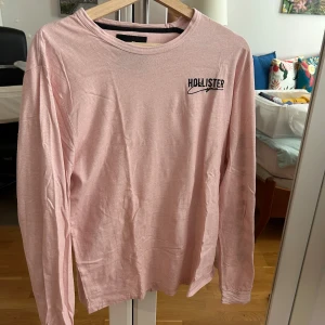 Ljusrosa långärmad tröja Hollister - Ljusrosa långärmad tröja från Hollister i 100% bomull. Tröjan har svart Hollister-logga på bröstet och stora bokstäver längs ena ärmen. Perfekt basic med relaxed vibe och rund halsringning.