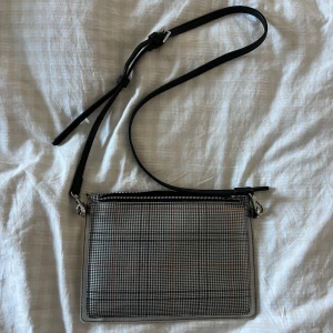 Rutig axelväska med dragkedja - Snygg och smidig axelväska med rutigt mönster i grått, svart och röda linjer. Väskan har en justerbar svart axelrem och öppnas med en silverfärgad dragkedja. Insidan har flera fack för kort och småsaker. Perfekt för att bära med det viktigaste.