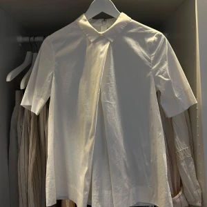 Vit skjorta med kort ärm från COS - Minimalistisk vit skjorta från COS i storlek 36, med korta ärmar och klassisk krage. Skjortan har en snygg asymmetrisk frontdetalj och stängs med en diskret dragkedja bak. Tillverkad i mjuk bomull som känns skön mot huden.