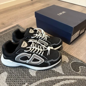Svarta Dior B30 sneakers med vita detaljer - Säljer ett par svarta Dior B30 sneakers med vita och silverfärgade detaljer. Skorna har en chunky vit och svart sula, tydlig CD-logga på sidan och snörning i vitt. Ovandelen är i mesh och syntet, vilket ger en sportig vibe. Perfekta för dig som gillar exklusiva sneakers med statement.