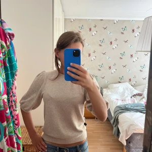 Beige puffärm stickad tröja 15A - Superfin beige stickad tröja från 15A med puffärmar och rund hals. Tröjan har en mjuk känsla och är perfekt för en lite svalare dag. Passformen är normal och ärmarna är trekvarts. Enkel att styla med jeans eller kjol. Den är nopprig men syns inte lika mycket i verkligheten. 