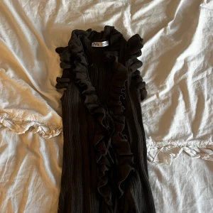 Brun volang topp från Zara - Säljer en brun ribbad tooo från Zara med snygga volangdetaljer längs axlar och framkant. Toppen är ärmlös och har en cool omlott-look framtill. Perfekt för dig som gillar trendiga och unika plagg med extra edge. Köpt ny på Plick men har inte kommit till användning 