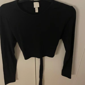 Svart croppad långärmad topp H&M - Svart långärmad topp från H&M med croppad passform och snygg knytning framtill i midjan. Enkel och stilren design med rund halsringning, perfekt att matcha med höga jeans eller kjol. Mjuk och skön bomullskvalitet.