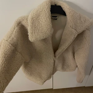 Beige teddyjacka med dragkedja - Mysig beige teddyjacka med stor krage och dragkedja framtill. Jackan har en fluffig och mjuk känsla, perfekt för kyliga dagar. Passformen är normal och modellen är klassisk med lång ärm. Enkel att matcha med både jeans och sneakers.