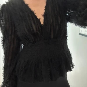 Svart transparent blus med struktur - Säljer en svart blus med transparent tyg och fluffig struktur. Djup v-ringning och långa ärmar. Midjan är markerad och tyget har en cool, randig effekt med fransiga detaljer. 