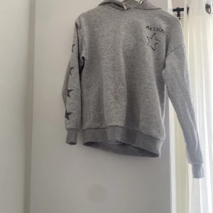 Grå hoodie med stjärnor och huva - Säljer en grå hoodie från Gina med tryckta svarta stjärnor längs ärmarna och en  stjärna med texten 'Ma Chérie' på bröstet. Tröjan har huva, ribbade muddar och är långärmad. Perfekt för chill dagar och enkel att matcha med jeans eller leggings.