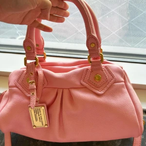 Rosa handväska från Marc by Marc Jacobs - Snygg rosa handväska från Marc by Marc Jacobs i mjukt skinn med guldfärgade metalldetaljer och dubbla handtag. Jag har aldrig använt den här väskan sen jag köpte den, och den är i superbra skick. Den är faktiskt ännu mer rosa i verkligheten än på bilderna. Ljuset inne när jag fotade var inte det bästa, så den ser ännu finare ut i verkligheten. Jag kan skicka fler bilder om du vill. Priset är redan riktigt bra med tanke på hur fint skick väskan är💕