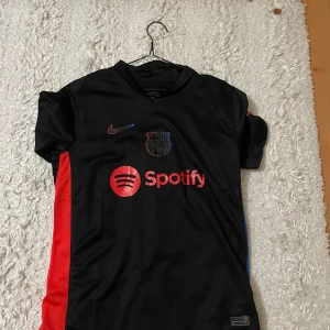 FC Barcelona Lamine Yamal tröja Nike - Snygg FC Barcelona matchtröja med Lamine Yamal och nummer 19 på ryggen. Svart bas med röda och blå detaljer, Spotify-logga på bröstet och UNHCR-tryck bak. Kortärmad och tillverkad i lätt, ventilerande material. Perfekt för dig som älskar Barca och vill ha en unik tröja.