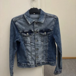 Klassisk blå jeansjacka med knappar - Snygg jeansjacka i blå denim med slitna detaljer och silverfärgade knappar framtill. Jackan har två bröstfickor med lock och knapp, klassisk krage och långa ärmar. Perfekt för dig som vill ha en tidlös och cool look.