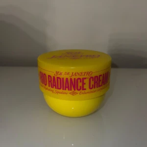 Sol de Janeiro Rio Radiance Cream - Superfräsch Rio Radiance Cream från Sol de Janeiro i en klargul rund burk med rosa text. Innehållet är en ljusbeige, krämig formula som ger glow och återfuktning med squalane. Perfekt för dig som vill ha en strålande finish på huden.