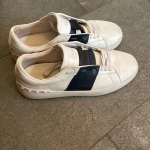 Valentino Garavani vita sneakers - Snygga vita Valentino Garavani sneakers med bred mörkblå rem över vristen och klassisk snörning. Skorna har rund tå och platt sula, tillverkade i skinn med diskret logga på plösen och innersulan. Perfekt för dig som gillar stilrena designer med en twist.