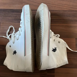 Vita high-top sneakers från Polo Ralph Lauren - Snygga vita high-top sneakers från Polo Ralph Lauren med klassisk snörning och rund tå. Skorna har en svart broderad logga på sidan och ränder i rött och grönt längs sulan. Ovandelen är i slitstark bomull och yttersulan är vit med transparent detalj under foten.