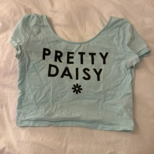 Ljusblå croppad topp - Superfin ljusblå croppad topp med svart texten 'PRETTY DAISY' och en liten blomma framtill. T-shirten har korta ärmar och rund halsringning. Perfekt för dig som gillar en söt och trendig stil.