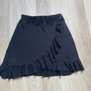 Svart volangkjol från Gina Tricot - Supersöt svart kortkjol från Gina Tricot i omlottmodell med volangdetaljer längs nederkant och framstycke. Kjolen har en enkel och stilren look som är lätt att matcha med olika toppar. Perfekt för dig som gillar trendiga plagg.